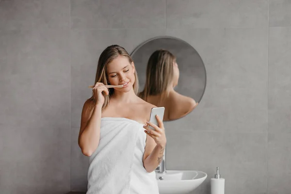 Pretty lady wrapped in towel using cellphone while doing morning routine in bathroom, brushing teeth — Stock Photo, Image