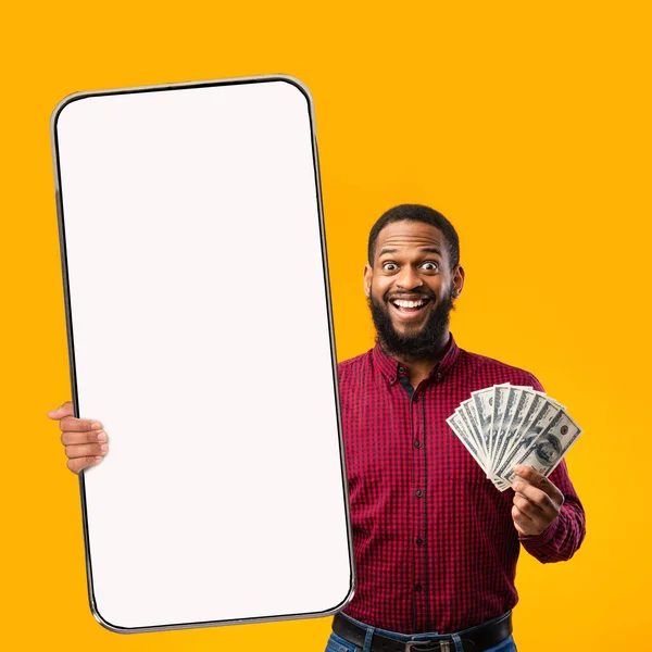 Glücklicher afrikanisch-amerikanischer Mann hält riesiges Smartphone mit leerem Bildschirm und Bargeld Dollar-Banknoten, Attrappe — Stockfoto