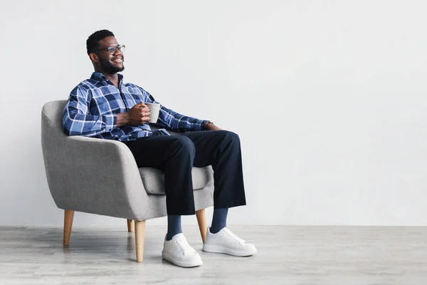 Descanso. Soñador joven afroamericano sentado en un sillón, disfrutando de una taza de bebida caliente contra la pared blanca — Foto de Stock