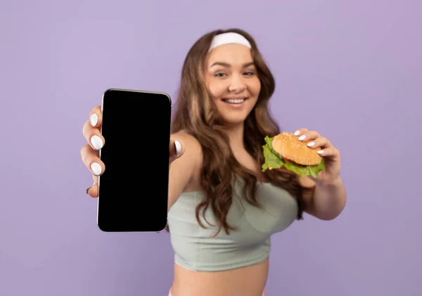 Χαρούμενη νέα συν μέγεθος γυναίκα σε αθλητικά ρούχα με burger δείχνει smartphone με λευκή οθόνη — Φωτογραφία Αρχείου