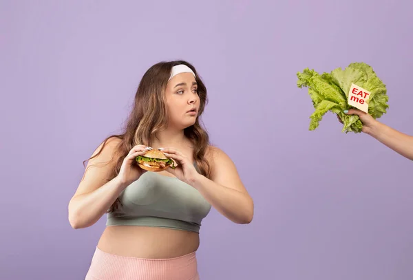 Zainteresowany głodny młody Europejczyk plus rozmiar pani w odzieży sportowej hold burger, spójrz na rękę z warzywami — Zdjęcie stockowe