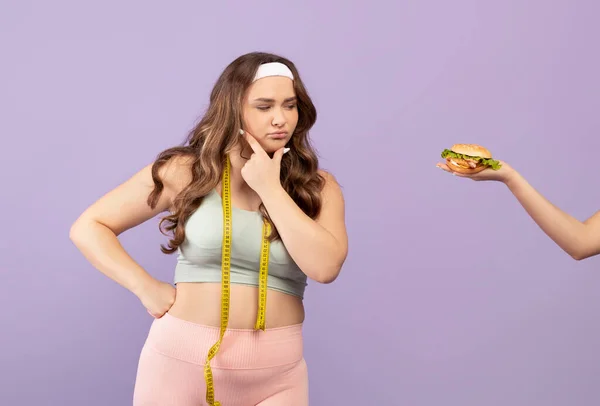 Pensive millennial caucasian plus size lady in sportswear with measuring tape looks at hand with burger — Stock Photo, Image