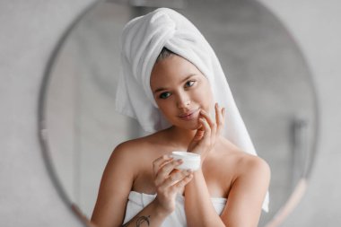 Güzel bir kadın yüz kremi kullanıyor, kavanozu tutuyor ve yanaklarına güzellik ürünü sürüyor, aynaya bakıyor.