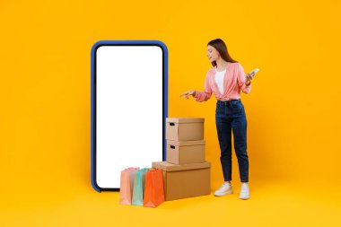 Büyük cep telefonlarını ve karton kutuları işaret eden kadın, sarı arka plan.