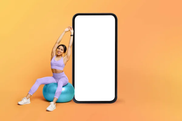 Aplicación de fitness. Atlético entrenamiento de señora negra en la pelota de fitness cerca de un gran teléfono celular con pantalla en blanco, maqueta —  Fotos de Stock