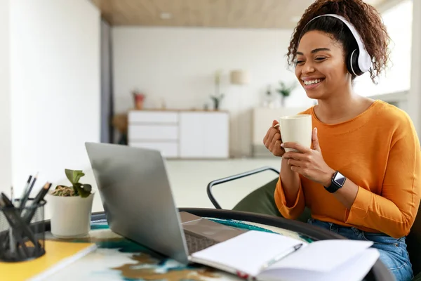 Uśmiechnięta czarna pani ogląda webinarium na komputerze, pije gorącą kawę — Zdjęcie stockowe