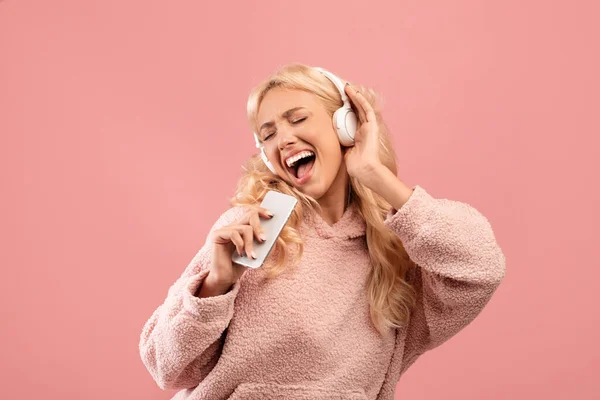 Hravý mladá dáma zpívá a drží smartphone jako mikrofon na sobě bezdrátové sluchátka, růžové pozadí — Stock fotografie