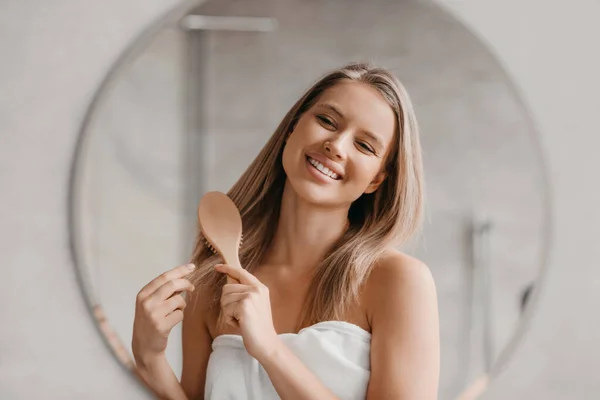 Концепція догляду за волоссям. Гарненька леді чистить її красиве волосся з гребінцем, дивлячись на її відображення у дзеркалі — стокове фото