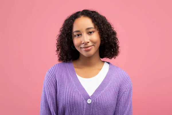Vrolijke jonge Afro-Amerikaanse vrouw met lichte glimlach en krullend haar op zoek naar camera over roze studio achtergrond — Stockfoto
