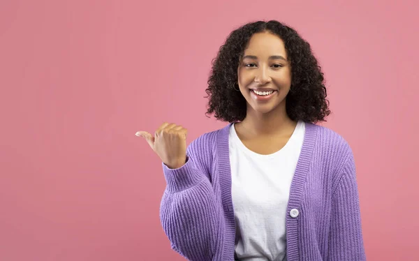 Joven mujer negra fresca en ropa casual señalando a un lado, ofreciendo espacio de copia para el diseño de su anuncio en el fondo del estudio rosa — Foto de Stock