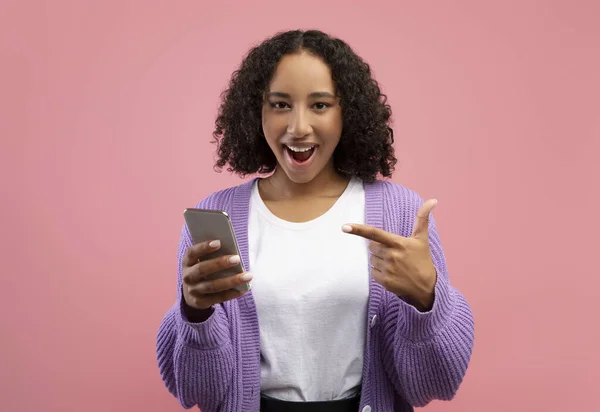 Emocionada joven mujer negra apuntando al teléfono inteligente, feliz por la enorme venta en línea en el fondo del estudio rosa — Foto de Stock