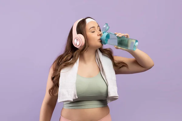 Törstig tusenårig europé plus storlek kvinna i sportkläder med handduk i hörlurar dricker vatten från flaska — Stockfoto