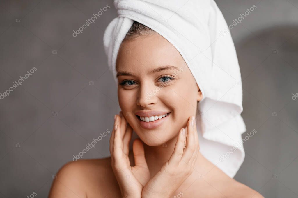 Happy lady posing with towel on head after bathing routine, standing in modern bathroom and smiling at camera