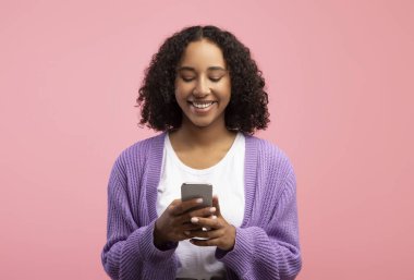 Akıllı telefon kullanan genç siyahi kadın portresi, sosyal medyada mesajlaşma veya pembe stüdyo arka planında görüntülü sohbet