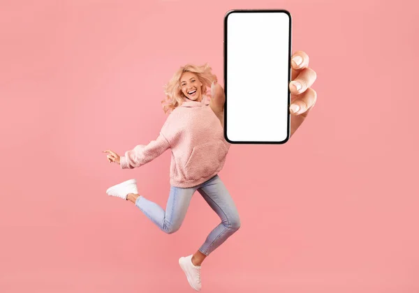 Возбужденная леди показывает пустой экран мобильного телефона во время прыжка на розовом фоне студии, коллаж, полная длина тела — стоковое фото
