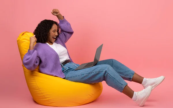 Jeune femme noire avec ordinateur portable levant les bras, célébrant le succès, assise dans une chaise de sac de haricots sur fond rose — Photo