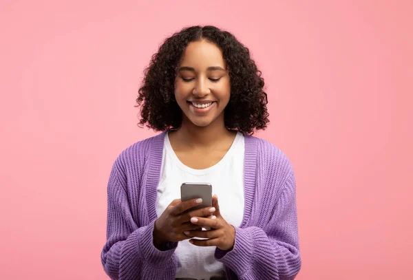 Portrét mladé černošky pomocí smartphone, zprávy v sociálních médiích nebo video chatování na růžovém pozadí studia — Stock fotografie