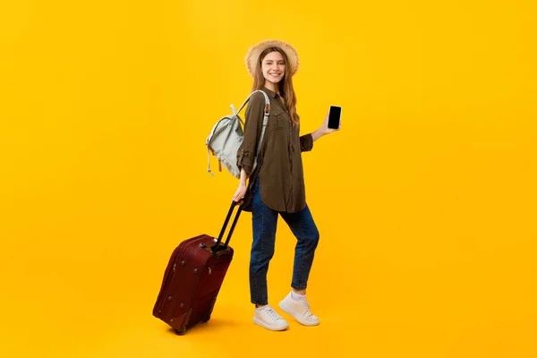 Turista femenina sosteniendo Smartphone posando con equipaje sobre fondo amarillo — Foto de Stock