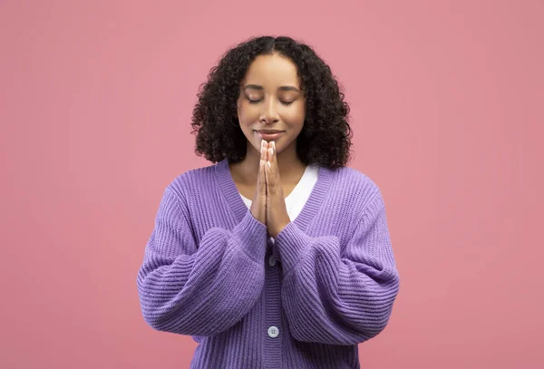 Мирная молодая афроамериканка молится Богу с сжатыми руками и закрытыми глазами на розовом фоне студии — стоковое фото