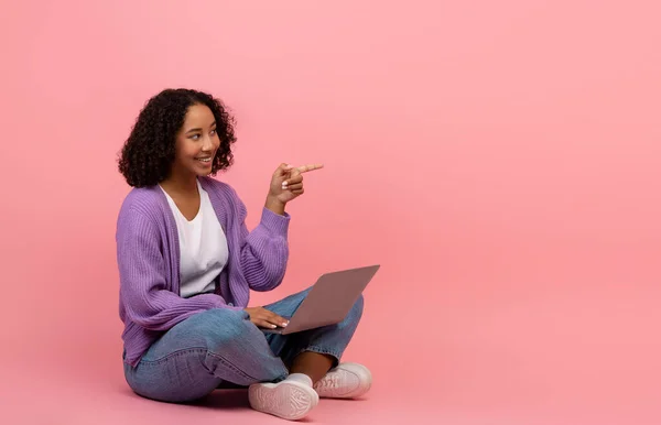かなり若いアフリカ系アメリカ人の女性がノートパソコンで足を組んで座って、ピンクの背景に空のスペースを指して — ストック写真