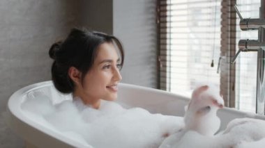 Rahatlamış kadın banyoda köpüklü banyo yapıyor, yandan bakıyor.
