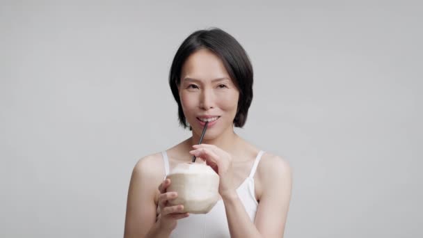 Gammal japansk kvinna dricker kokosvatten med halm, grå bakgrund — Stockvideo