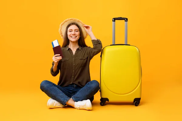 Mujer viajera feliz sosteniendo el pasaporte sentada cerca de la maleta, fondo amarillo — Foto de Stock
