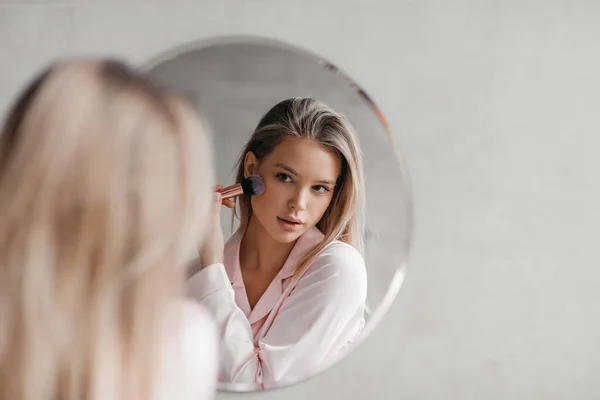 Mulher bonita em pijama na frente do espelho, segurando escova e aplicando maquiagem no banheiro, espaço livre — Fotografia de Stock