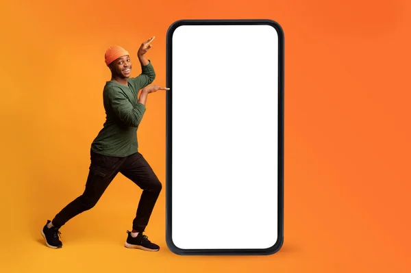 Komik Afrikalı Amerikalı adam büyük bir cep telefonunun yanında boş ekranla dans ediyor ve turuncu arka planda eğleniyor. — Stok fotoğraf