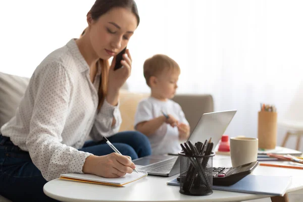 Junge, geschäftige Mutter arbeitet, während ihr kleiner Sohn neben ihr zeichnet, Frau telefoniert und Notizen macht — Stockfoto