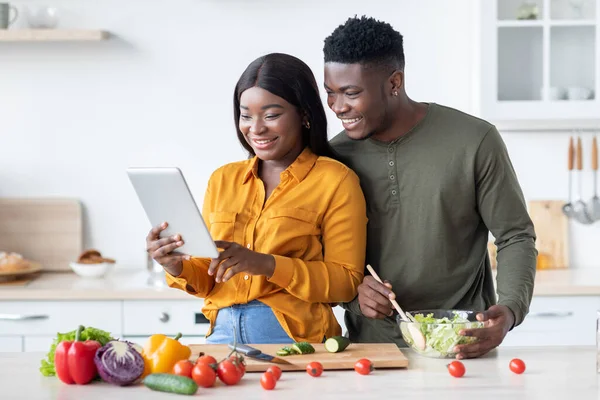 Cónyuges negros felices usando la tableta digital mientras cocinan el almuerzo en la cocina juntos — Foto de Stock