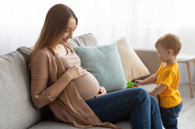 Mutlu hamile anne kanepede oturup karnına dokunuyor, küçük çocuğuyla evde vakit geçiriyor.