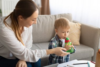 Doğum izni konsepti. Genç anne evde internetten çalışırken oğluna su şişesi veriyor.
