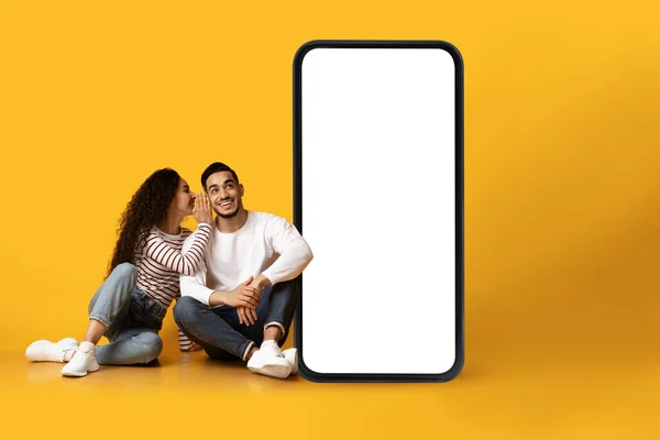 Молода арабська пара сидить біля смартфона з білим екраном, жінка розповідає чоловікові про круту пропозицію, сміється за дизайн — стокове фото