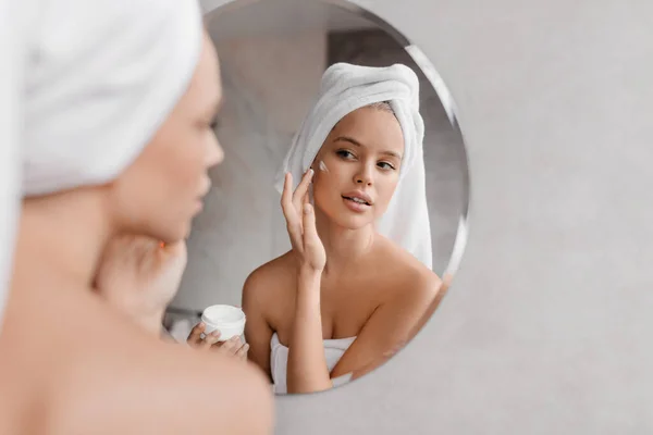 Home beauty routine. Young woman applying moisturising cream on face looking at miror in bathroom, free space — Stock Photo, Image