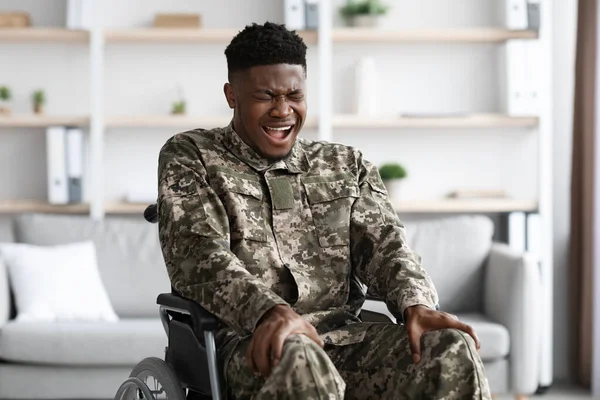 Boldogtalan, tolószékes fekete katona, aki a lábát masszírozza és sikoltozik. — Stock Fotó