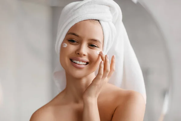 Cabeza de mujer aplicando crema facial en las mejillas, sonriendo a su reflejo en el espejo, disfrutando de la rutina de belleza —  Fotos de Stock