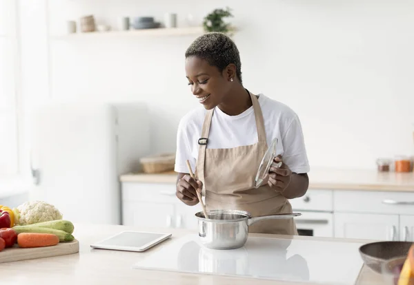 앞치마를 입은 아프리카 계 미국인 아가씨가 요리를 하고 태블릿 동영상 강의를 보고 있어서 기쁩니다. — 스톡 사진