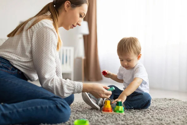 Молодая мама и маленький ребенок играют вместе дома, используя игрушки, сидя на полу ковра в гостиной — стоковое фото