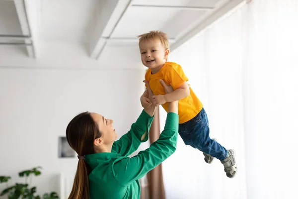 Moederschap concept. Speelse dame houdt haar lachende kind jongen, tillen zoon hoog in de lucht, plezier hebben thuis — Stockfoto