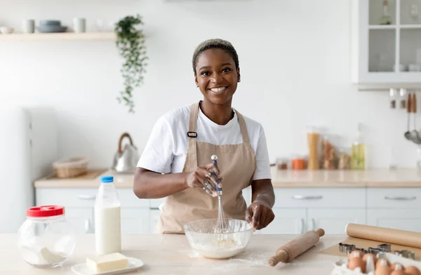 Smiling pretty millennial african american woman chef in apron making dough in light minimalist kitchen interior — Stock Photo, Image