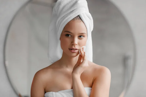 Ρουτίνα ομορφιάς. Όμορφη νεαρή γυναίκα καθαρίζει το πρόσωπό της με βαμβάκι στο σπίτι, στέκεται με πετσέτα στο κεφάλι μετά το μπάνιο — Φωτογραφία Αρχείου