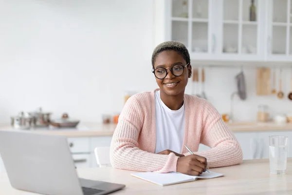 Glad millennial afroamericana mujer en gafas de trabajo o estudio en el ordenador portátil en el lugar de trabajo en el interior de la cocina —  Fotos de Stock