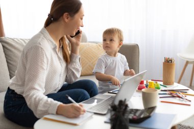 Evde çocukla çalışan, cep telefonuyla konuşan ve not defterine not alan meşgul genç bir anne.