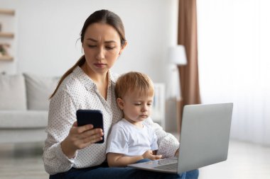 Genç anne akıllı telefon kullanıyor, dizüstü bilgisayarda çalışırken kötü haberler okuyor dizlerinin üstüne çöküp uzayı kopyalıyor.