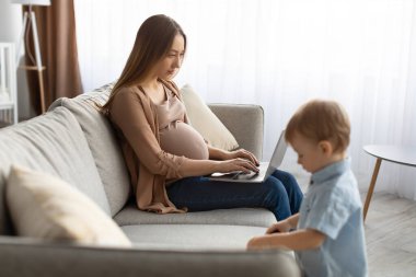 Küçük oğluyla evde vakit geçirirken dizüstü bilgisayarda çalışan hamile bir kadın.