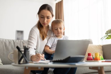 Mesafe öğrenme. Çocuklu genç bayan internetten ders çalışıyor, web sitesi izliyor ve not defterine notlar yazıyor.