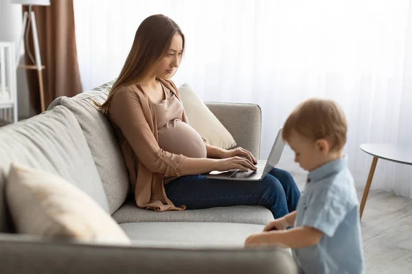 Jeune femme enceinte travaillant sur un ordinateur portable tout en passant du temps avec son petit fils à la maison — Photo