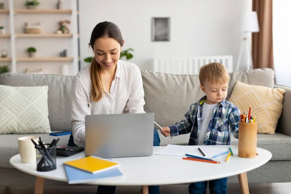 Молодая мать работает онлайн на ноутбуке с сыном играть вокруг, леди фрилансеров и заботиться о своем ребенке малыша — стоковое фото