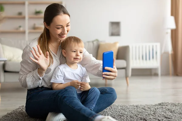 Jovem mãe branca e pequeno filho usando smartphone e fazendo videochamada, mulher acenando olá para o celular — Fotografia de Stock